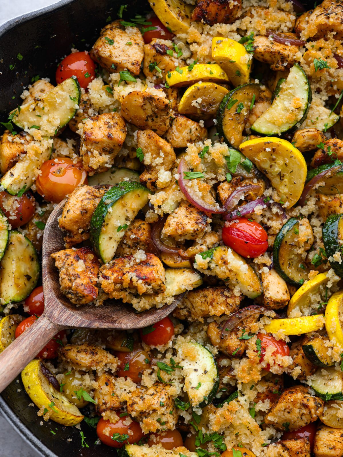 Use up all those garden veggies in this delicious chicken and garden vegetable skillet. Your family will love this healthy 30-minute skillet dinner full of seasoned chicken, sauteed vegetables, and a cheesy breadcrumb topping. Close up view of a wooden spoon lifting up the chicken and garden vegetables from the skillet.