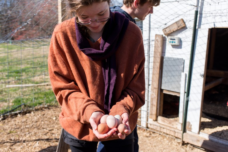 A young person in an orange sweater and purple scarf holds eggs in shades from white to brown in two hands, smiling while looking down at them. A person stands behind her, looking at a hen house.